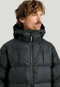 Mustat untuvatakki, jossa on tikattu muotoilu, korkea kaulus, säädettävät nyörit ja etuvetoketju. Sileä pinta, minimaalinen brändäys.