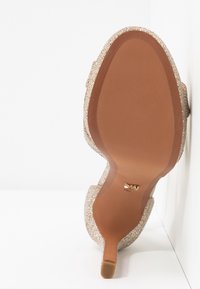 Vue de la semelle d'une chaussure à talon haut pailletée avec un talon étroit et une semelle extérieure lisse de couleur marron sur fond blanc.