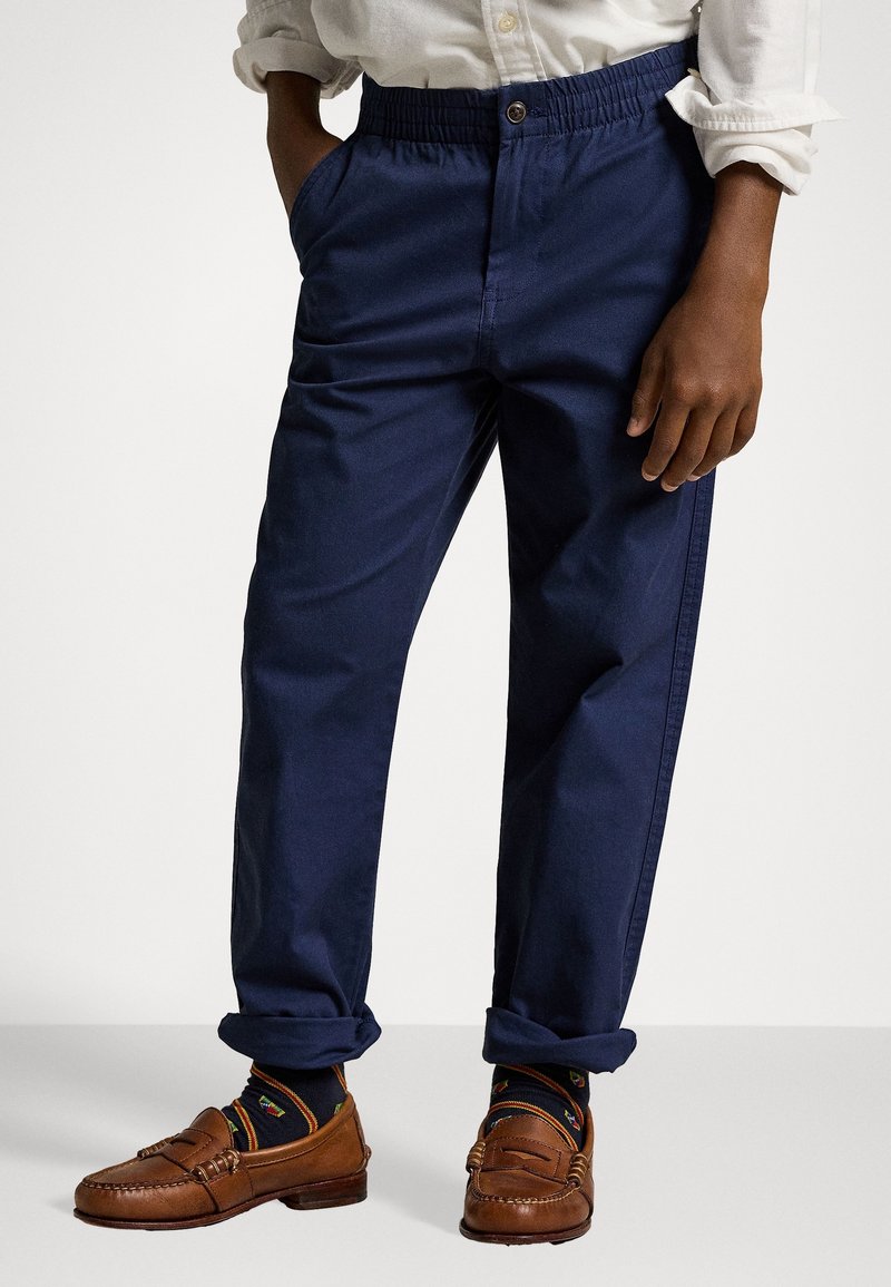 Personne portant un pantalon bleu marine à taille élastique, des chaussettes foncées à motifs, des mocassins en cuir marron et une chemise blanche à manches longues retroussées.