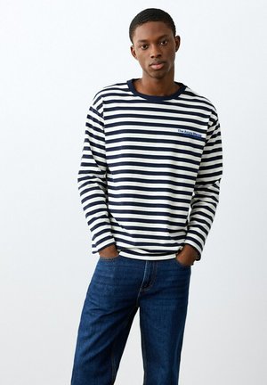 Giovane uomo che indossa una maglia a maniche lunghe a righe blu navy e bianche con scritta e jeans blu, in piedi con le mani in tasca davanti a uno sfondo bianco.