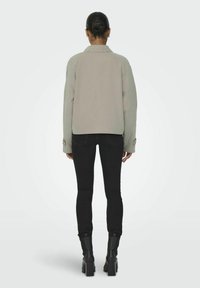 Chaqueta gris claro con un ajuste ligeramente estructurado, que cuenta con mangas largas y puños abotonados, combinada con jeans negros ajustados y botas hasta el tobillo.