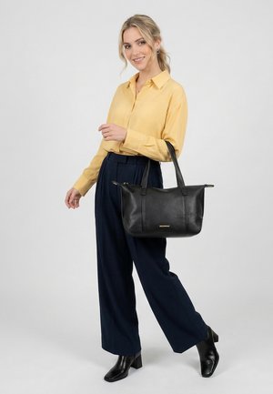 Donna che indossa una camicetta gialla, pantaloni larghi blu navy, stivaletti neri e tiene una borsa in pelle nera con scritte dorate.