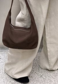 Brun mockaläder axelväska med en böjd form och en silverfärgad dragkedja på framsidan, kombinerad med löst sittande beige byxor och svarta skor.