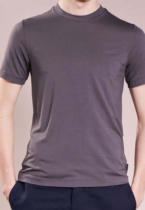 Homme portant un t-shirt ajusté à manches courtes gris foncé et un pantalon bleu marine, se tenant avec les mains dans les poches devant un fond neutre.