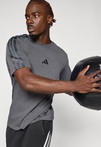 T-shirt de sport gris à manches courtes, avec trois rayures sombres sur les épaules et un logo sur la poitrine. Ballon de fitness noir tenu à la main.