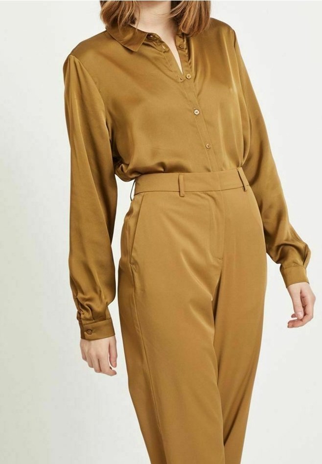 Femme portant un chemisier jaune moutarde brillant à manches longues, rentré dans un pantalon jaune moutarde taille haute avec poches avant.