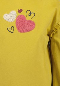 Maglietta gialla a maniche lunghe conApplicazioni a forma di cuore in tessuto rosa, bianco e bordeaux, con contorni neri e dettagli arricciati sulle maniche.