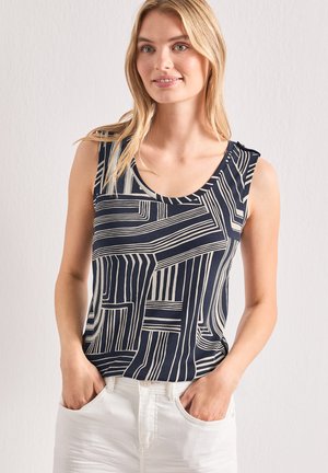 Femme portant un haut sans manches bleu marine avec des lignes géométriques beiges et un pantalon blanc, debout les mains dans les poches contre un mur uni.