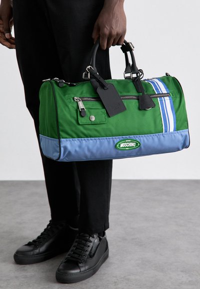 Vihreä ja sininen nahka-asusteella varustettu nailonista valmistettu duffel-laukku, jossa on yläkahvat, vetoketjulliset taskut ja näkyvä Moschino-logo.