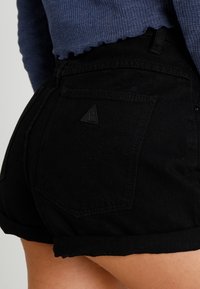 Personne portant un short en denim noir avec un petit logo triangulaire sur la poche arrière et un haut à manches longues côtelé bleu.