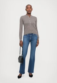 Cardigan grigio a coste con chiusura a bottoni, abbinato a jeans svasati blu. Il modello tiene in mano una piccola borsa nera con texture e indossa delle ballerine nere.