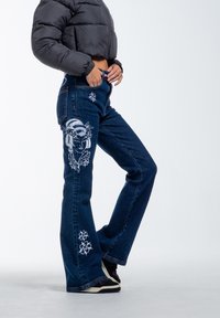Dark blue denim flared jeans with white embroidery of a face and flowers on the thigh and lower leg. High-waisted design, casual fit.