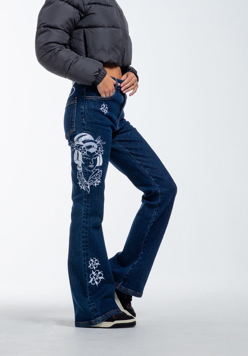 Dark blue denim flared jeans with white embroidery of a face and flowers on the thigh and lower leg. High-waisted design, casual fit.