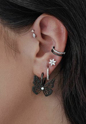Black metal butterfly earring with intricate cutouts and a central rhinestone, alongside silver studs and hoops on the ear.