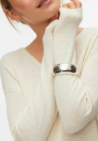 Femme portant un pull en tricot crème à manches longues et un large bracelet manchette en argent à son poignet près du visage.