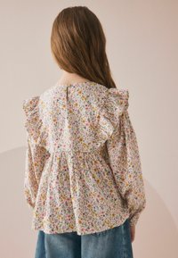 Blumenbluse mit Rüschenschultern, gerafftem Taillenbereich und Knopfverschluss hinten. Der Stoff zeigt ein mehrfarbiges Blütenmuster auf hellen Grund.