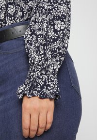 Blusa floreale con sfondo blu navale, caratterizzata da un motivo floreale bianco. Maniche a sbuffo e una vestibilità comoda, abbinata a denim scuro. Cintura nera visibile.