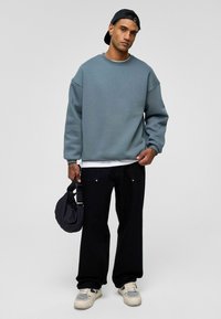 Blauwe sweatshirt met verlaagde schouders, zwarte wijde broek en beige sneakers. Houdt een zwarte tas vast, staat tegen een effen grijze achtergrond.
