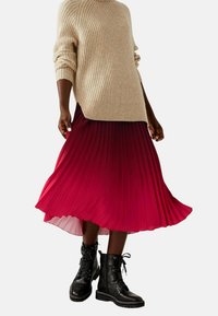 Beiger, gerippter Pullover, knie langer tiefrosa plissierter Rock und schwarze, geschnürte Stiefeletten. Der Rock zeigt einen Farbverlauf und Bewegung.