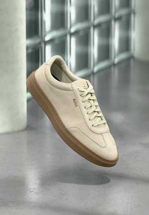 Jasnobeżowy zamszowy sneaker z gumową podeszwą w kolorze karmelu, wyrazistymi sznurowadłami i subtelnymi detalami przeszyć. Czysty design o minimalistycznej estetyce.