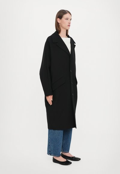 Svartullskappa med överdimensionerad passform, stora slag, tre knappar och två sidofickor, i kombination med blå vida jeans och svarta ballerinas.