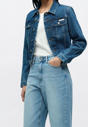 Person iført en blå denimjakke over en hvid top med den ene hånd i lommen på højhøj talje lyseblå jeans mod en hvid baggrund.