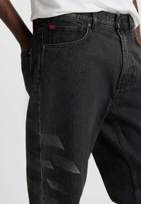 Black denim jeans featuring a faded textured pattern on the side, red logo tag, and contrasting stitching along the pockets and waistband.