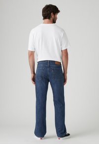 Jeans de mezclilla azul con corte recto, bolsillos traseros y una etiqueta de marca de cuero marrón; combinados con una camiseta blanca de manga corta.