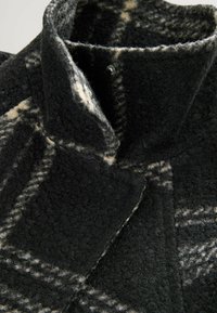 Manteau en laine à carreaux noir et beige avec un col haut et une fermeture à boutons-pression, montrant une texture détaillée et des plis du tissu.