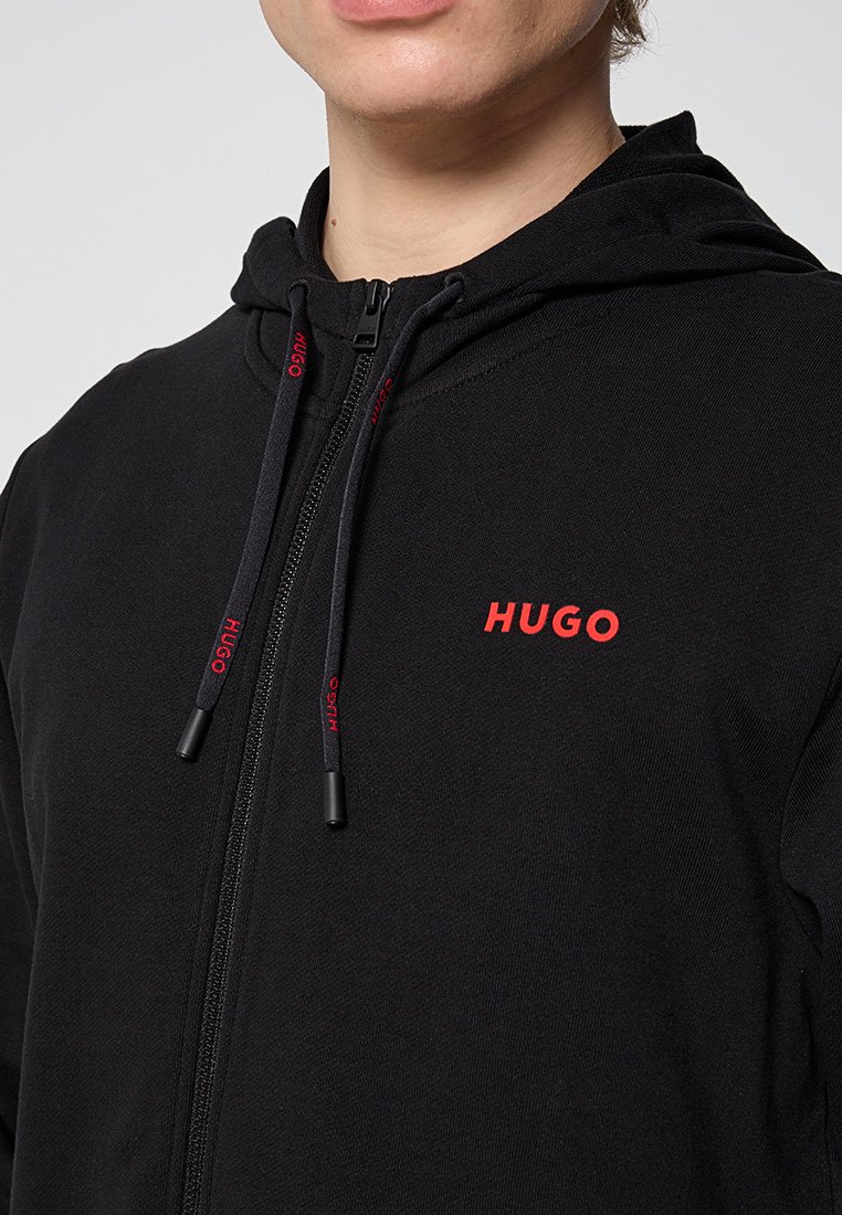 Schwarze Zip-up-Hoodie aus glattem Stoff, mit einem vorderen Reißverschluss, verstellbarem Kordelzug und einem roten "HUGO"-Logo auf der Brust.