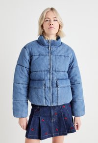 Denim puffer jacket with a high collar, front zip closure, and two large pockets. Light blue color, featuring horizontal stitching details.