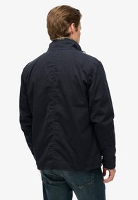 Marineblauwe jas met een hoge kraag, stoftextuur en knoopdetails bij de manchetten. Bevat een ruitvoering die zichtbaar is bij de kraag.