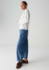 Donna in piedi di lato che indossa un maglione bianco a maniche lunghe, jeans blu larghi e sneakers marroni su uno sfondo chiaro e uniforme.