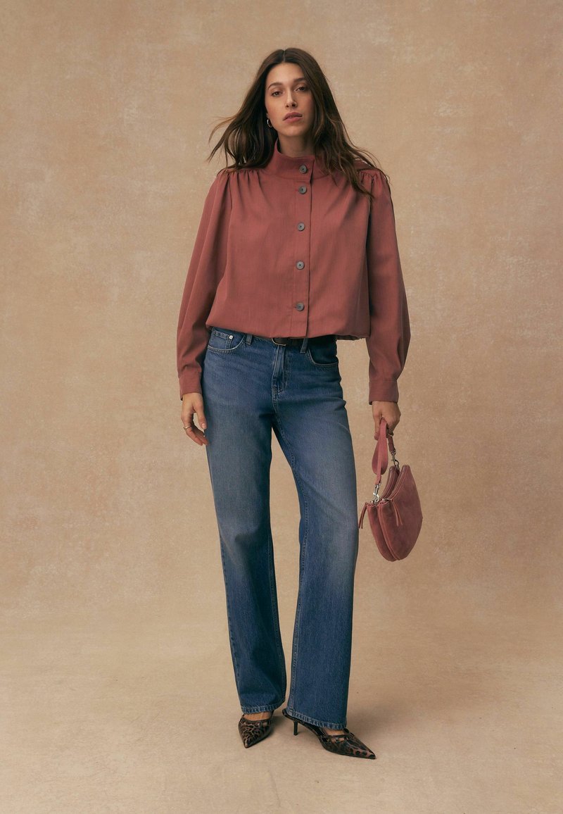 Frau trägt eine rostfarbene, geknöpfte Bluse, blaue Jeans, Leopardenmuster-Absätze und hält eine passende rostfarbene Handtasche.