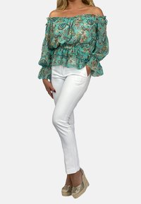 Off-shoulder turkos blus med paisleymönster, volangärmar, i kombination med vita figurnära byxor och beiga espadrillos.