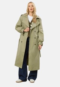 Oliwkowy płaszcz trench z podwójnym zapięciem, dużymi klapami i mankietami z paskiem. Noszony na ciemnozielonym swetrze i szerokich dżinsach.