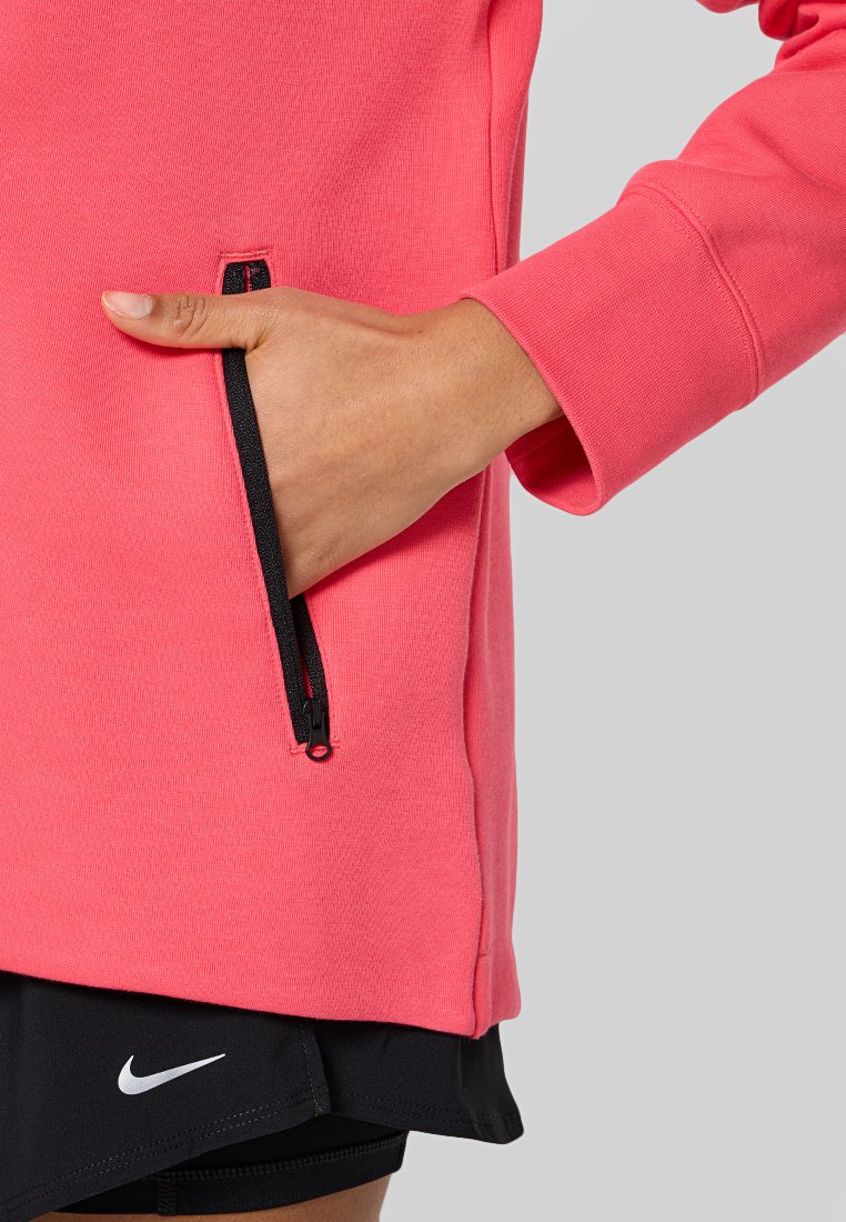 Sudadera rosa con bolsillo de cremallera negra y textura acanalada, con un tejido suave. Llevada con pantalones cortos negros que muestran un logo.