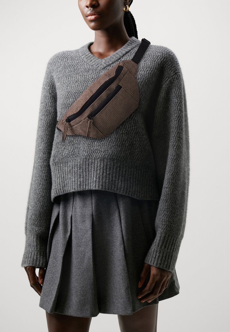 Ribbad brun crossbody-väska med två dragkedjefickor, bärs över en grå stickad tröja och en veckad kjol. Texturerad tyg, kompakt form.