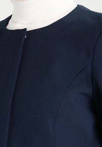 Cappotto blu scuro con colletto arrotondato, tessuto morbido e dettagli di cucitura discreti, indossato sopra un dolcevita bianco a coste.