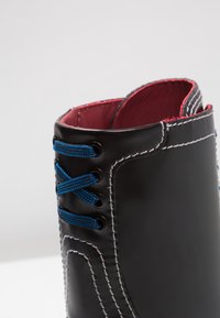 Gros plan d'une chaussure en cuir noire avec des coutures blanches, des lacets bleus et une doublure intérieure rouge.