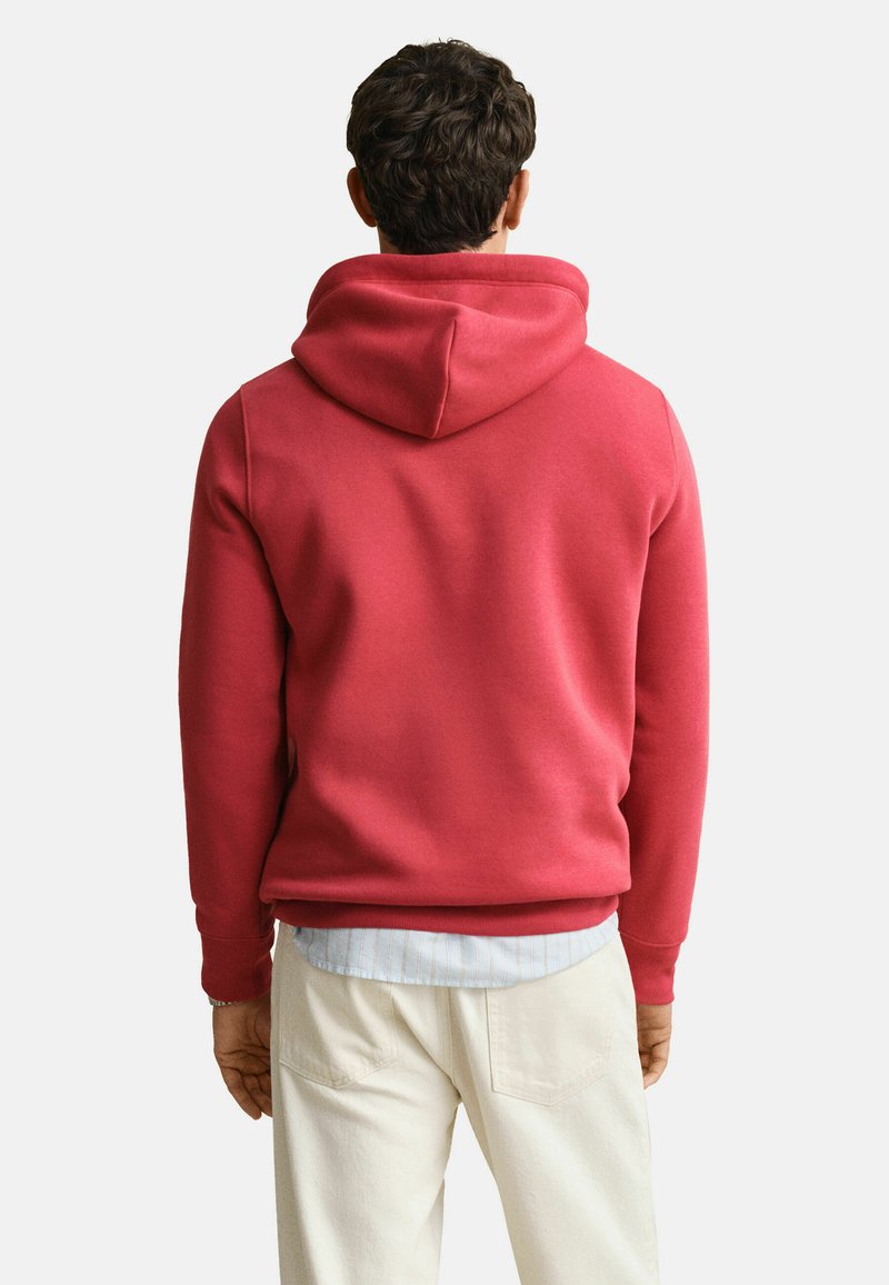 Ein Mann trägt einen roten Kapuzenpullover über einem hellblauen Hemd und cremefarbenen Hosen und steht mit dem Rücken zu einer schlichten Wand.