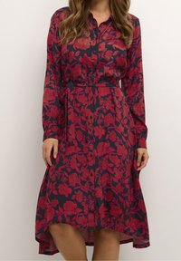 Donna che indossa un vestito lungo con maniche lunghe, abbottonato, con un motivo floreale rosso su sfondo scuro, con vita con cintura e orlo irregolare.