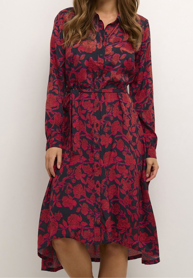 Donna che indossa un vestito lungo con maniche lunghe, abbottonato, con un motivo floreale rosso su sfondo scuro, con vita con cintura e orlo irregolare.