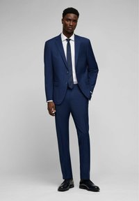 Completo blu navy con due bottoni, rever a punta e vestibilità su misura. Camicia bianca e cravatta abbinata, completato con scarpe eleganti nere.