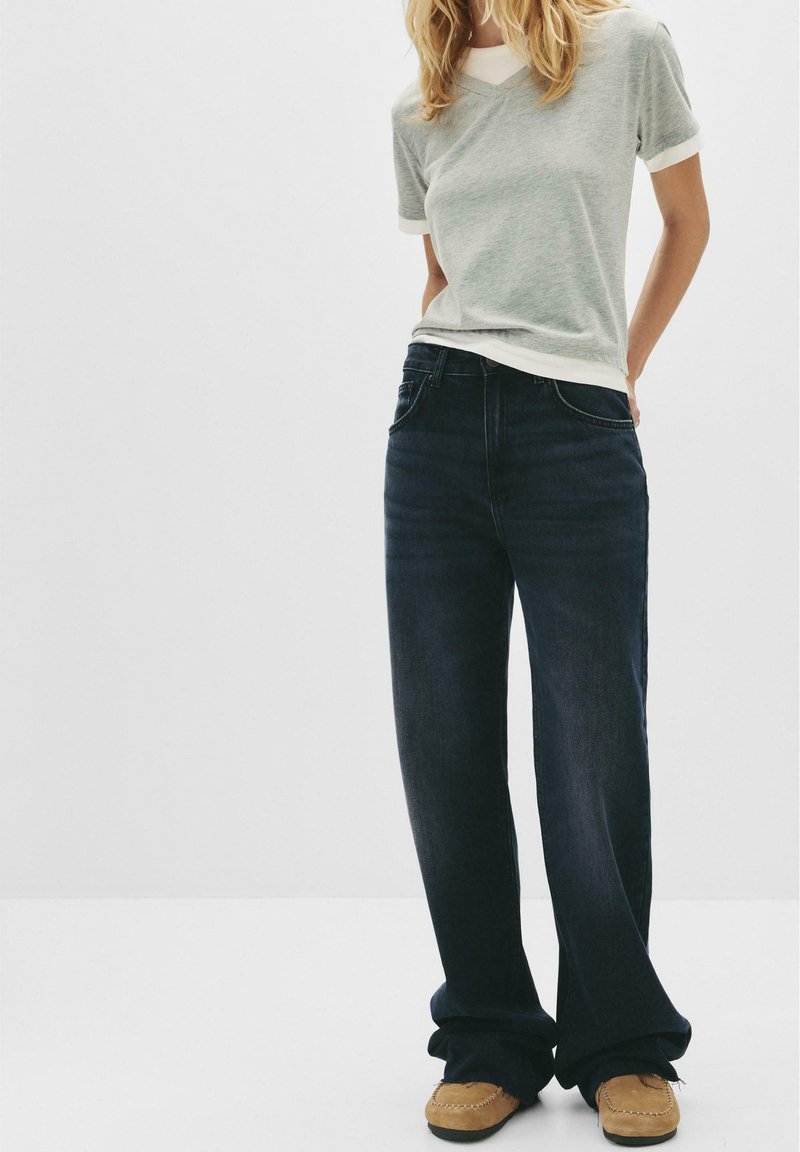 Mörkblå vida jeans i bomullsblandning med en slät textur. I kombination med en ljusgrå t-shirt med v-neck och vita kantdetaljer. Bruna mokasins.