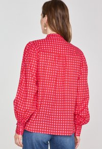 Blusa rossa con motivo a griglia bianca, maniche lunghe a sbuffo e chiusura con bottoni sul retro. Tessuto leggero con una vestibilità comoda.