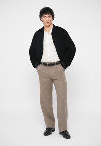 Άνδρας που στέκεται με τα χέρια στις τσέπες, φορά μαύρη ζακέτα, λευκό πουκάμισο με κουμπιά, μπεζ παντελόνι, μαύρη ζώνη και μαύρα παπούτσια σε λευκό φόντο.