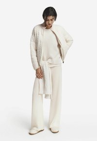 Pull en laine blanche à maille torsadée, pantalon large assorti et blouson aviateur matelassé. Texture douce, coupe décontractée et look superposé avec une écharpe.