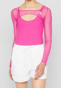 Body in lycra rosa con maniche lunghe trasparenti, caratterizzato da un design con ritaglio e accento con logo. Abbinato a pantaloni corti bianchi con cordoncino.