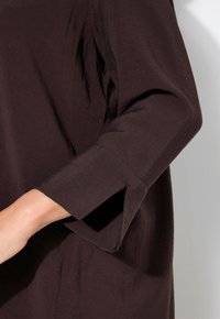 Donkerbruin langhoudend shirt met een ronde halslijn en een split bij de manchetten. De stof lijkt zacht en lichtjes gestructureerd.
